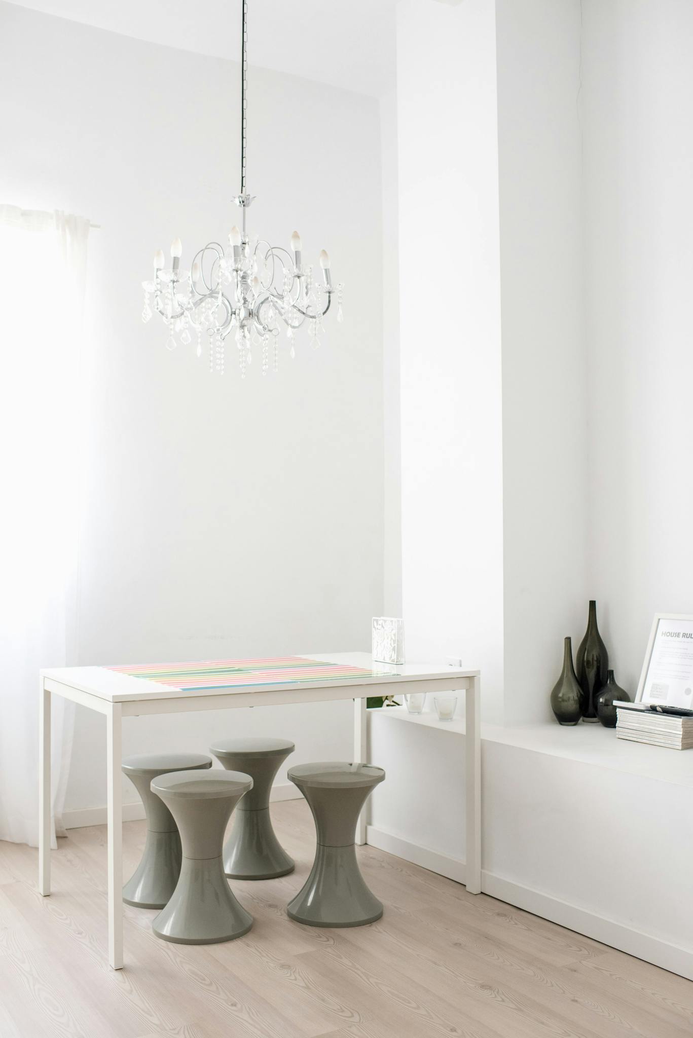 White table with chairs of unique shape placed near wall niche with black ceramic vases under crystal vintage chandelier in spacious light room with white walls and wooden floor